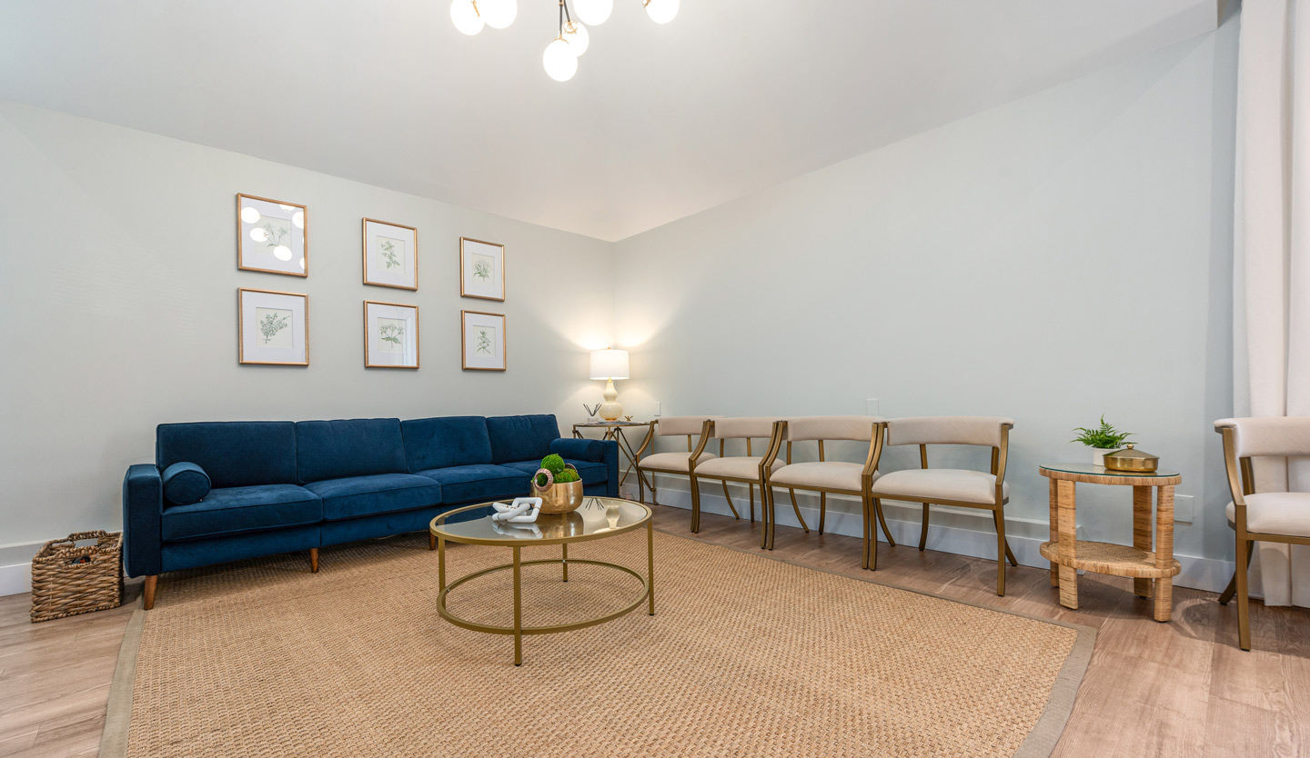 Twickenham Family Dentistry A modern waiting room with a navy blue couch, beige chairs, and a glass coffee table on a woven rug. The walls are adorned with framed botanical prints, and soft lighting adds warmth.